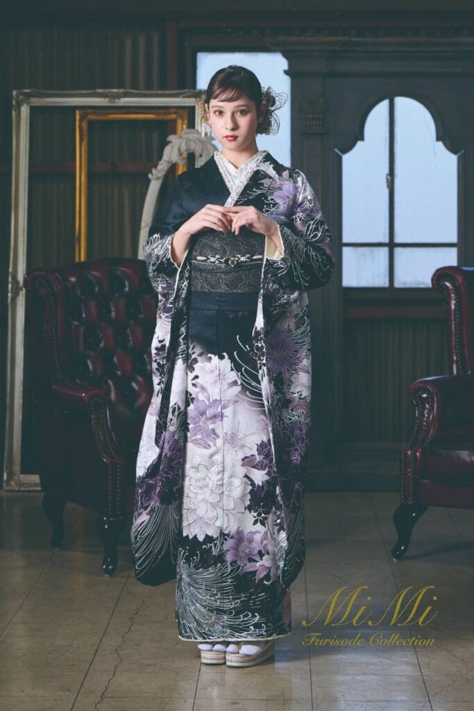 室内で、白と黒の花柄の着物を着て、濃い色の帯を締めた女性が、2脚の革張りの椅子と、装飾の施された空の額縁の隣に立っている。.