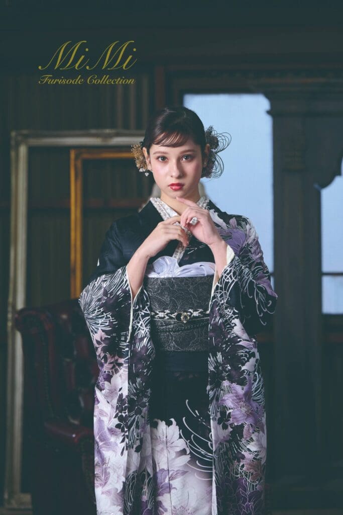黒とラベンダー色の花柄の着物を着た女性が屋内に立ち、カメラの方を見つめている。彼女の上には「Mi Mi 振袖コレクション」という文字が書かれている。.