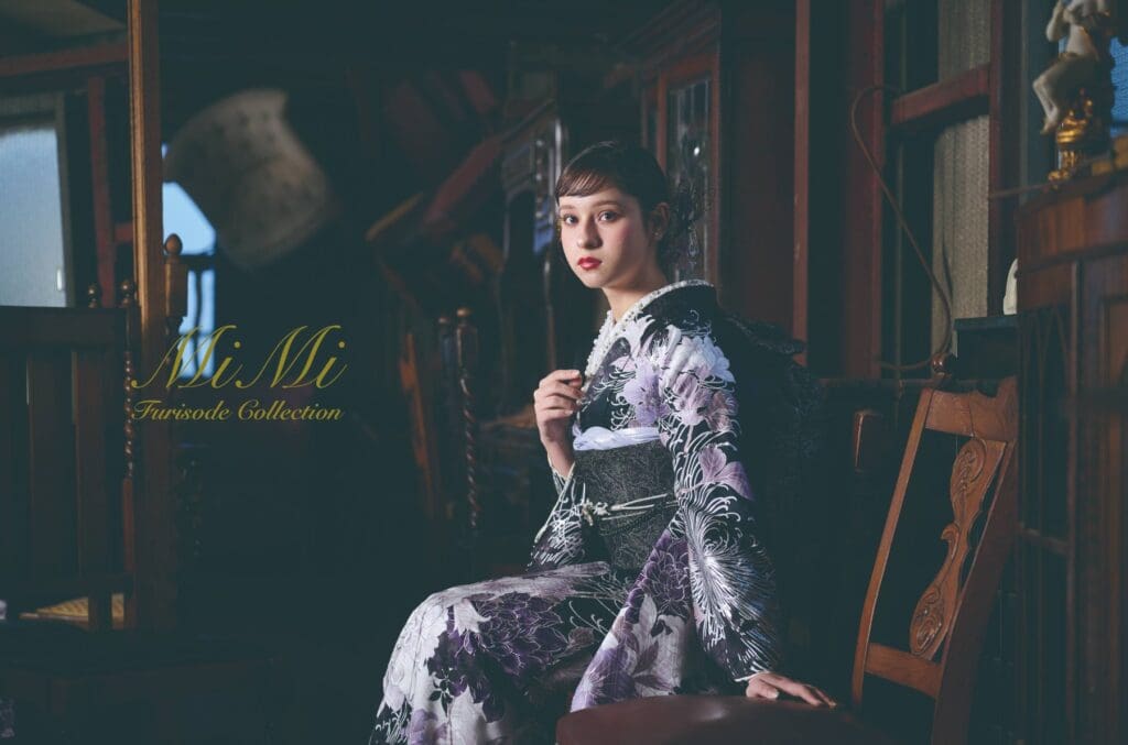 柄入りの着物を着た女性が、屋内の木製の椅子に座っている。画像の左側には「MiMi 振袖コレクション」という文字が表示されている。.