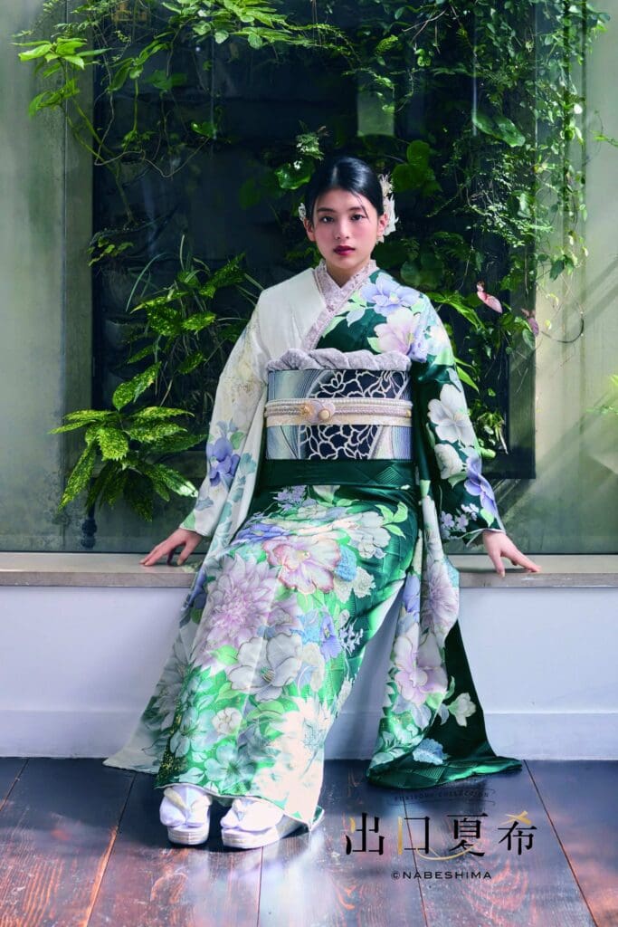 緑と白の花柄の着物を着た女性が、植物や緑を背景に、窓枠の上に座っている。.