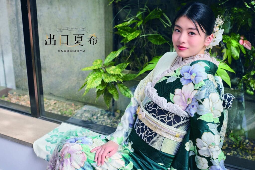 緑と白の花柄の着物を着た女性が、室内の窓辺に座っており、背後には緑の植物が見える。.