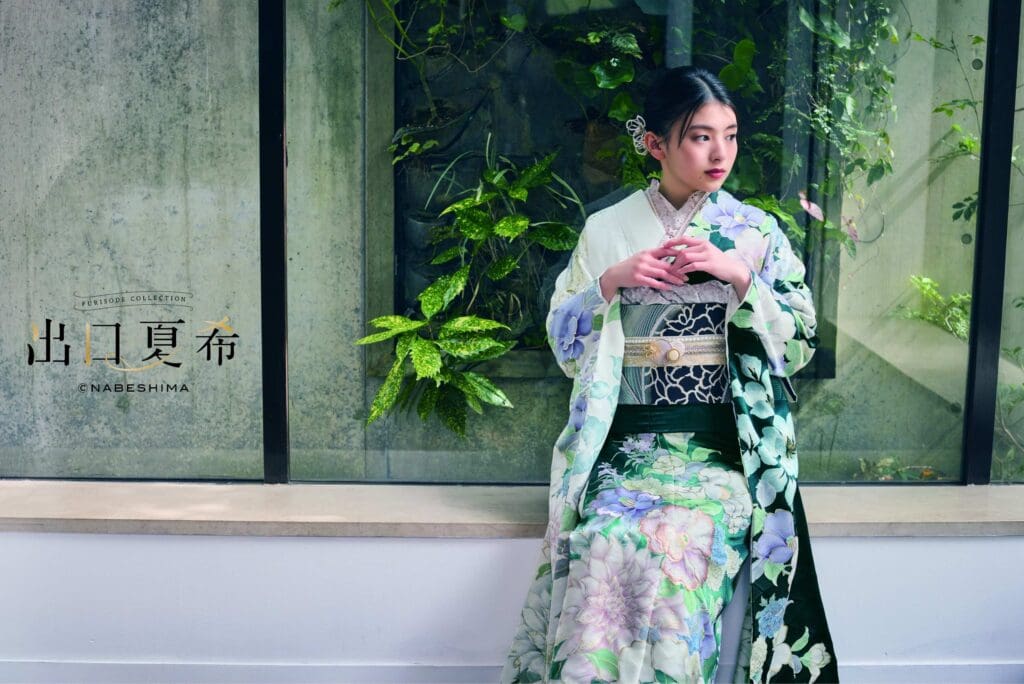 花柄の着物を着た女性が、屋内のコンクリートの縁に腰を下ろしており、大きな窓の向こうには青々とした植物が見える。.